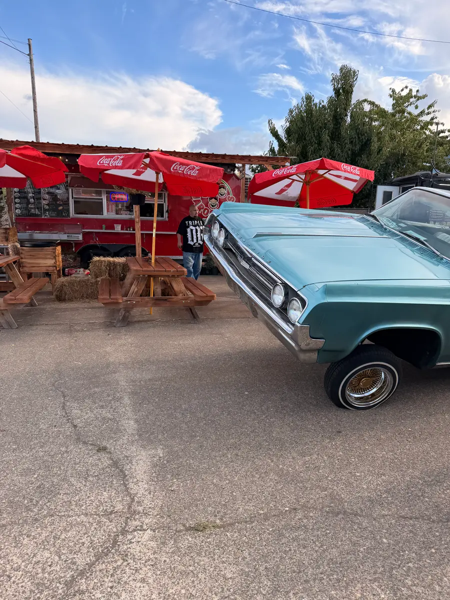 A teal lowrider with its front end raised, beside red umbrellas, picnic tables and a food truck at the park